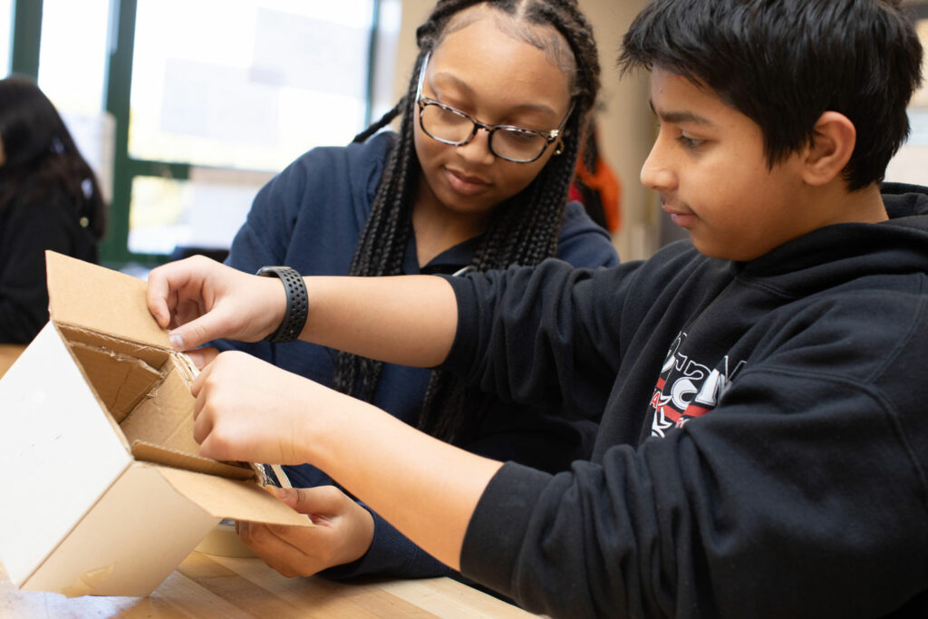 Two students construct a project with a cardboard box